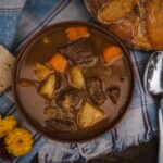 Beef stew with carrots and potatoes in brown ceramic bowl, served with bread