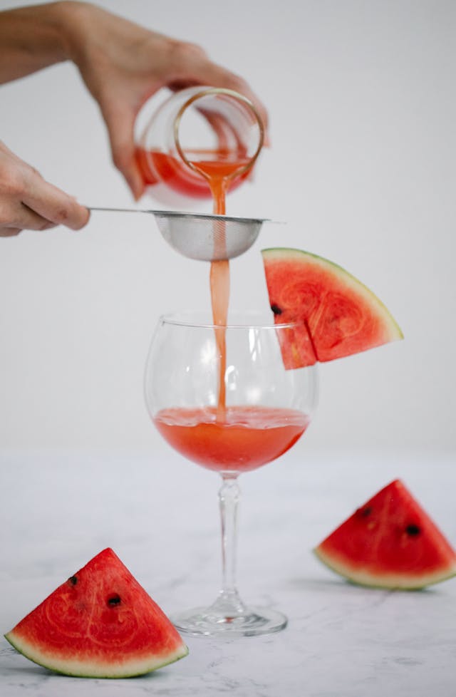 A bartender using a strainer to pour watermelon mint mojito mocktail from a shaker into a clear glass, ice and mint leaves visible, action shot, professional lighting.